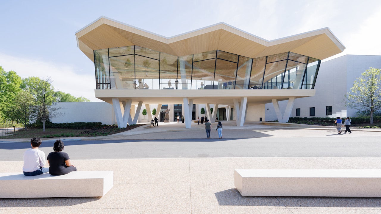 An outdoor setting that shows the façade of a contemporary building. A large walkway with white concrete, block-style benches is in the foreground. The building is raised more than 12 feet off the ground and sits on V-shaped stilts. The front of the building is an all-glass façade and people can be seen inside. The roofline is in a zig-zag shape. The colors are all white and cream with black window panes.