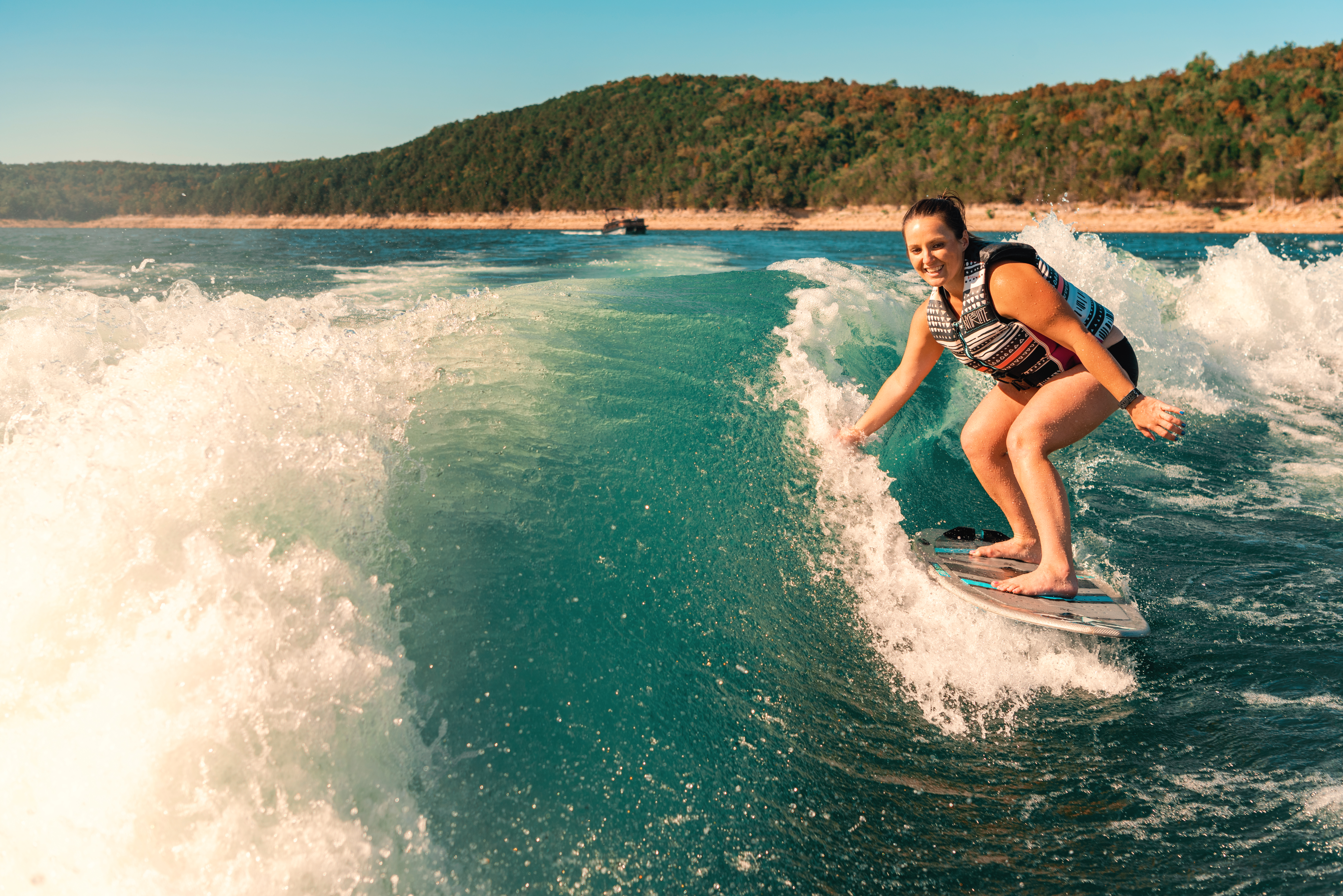 A lake setting near Mountain Home, Arkansas. Blue sky and green-tree hills are seen in the background. Blue water and white wake fill the rest of the photograph. A girl is seen on a board surfing in the wake. Photo source: Magnetic Creative.