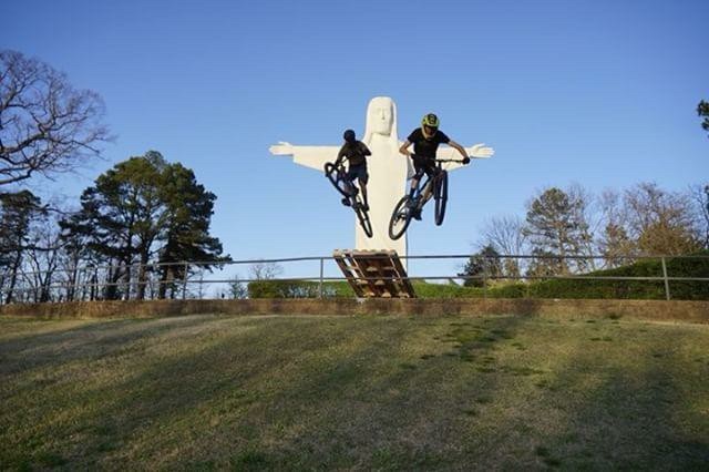 An outdoor setting with blue skies and green grass. A large white statue of Christ with his arms outstretched sits in the distance. In the foreground, two mountain bikers are ramping off a jump. 