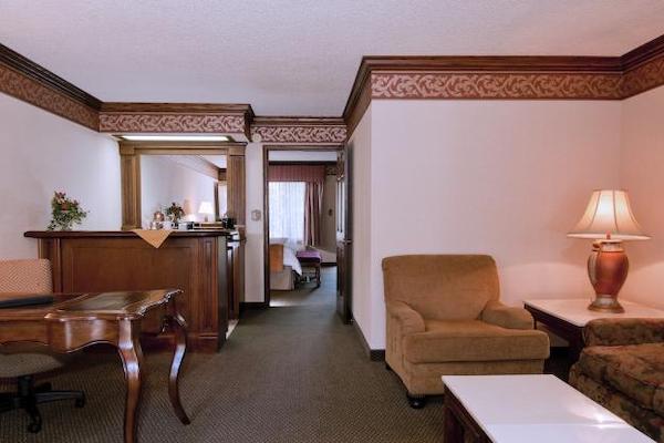 A hotel room. The first space is a living area with tables, a seating area and a wet bar. Through a doorway, a separate bedroom can be seen in the distance. 