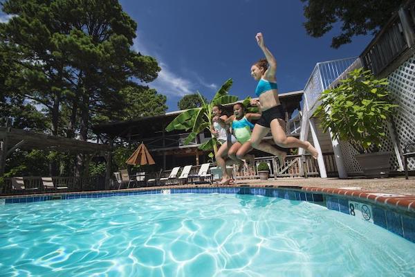 The edge of a pool with blue water. A group of three girls holding hands are mid-air in a jump into the pool from a concrete side. 