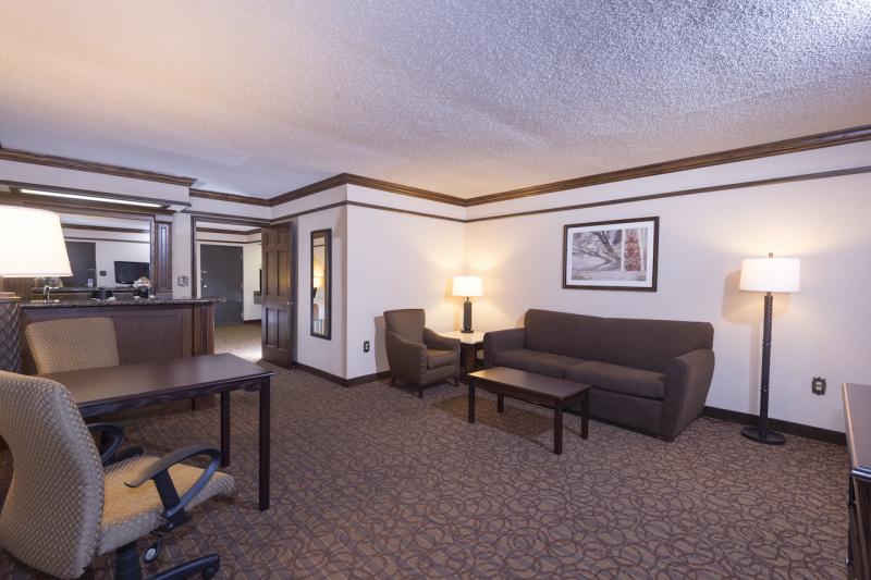 A hotel room shows a living area with a sofa, chair and coffee table. Opposite this conversation area is a desk and desk chair along with a wet bar against a back wall. 