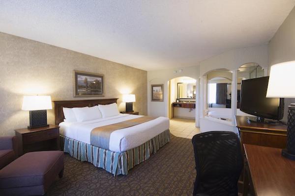 A hotel room. A king-size bed is in the center. A desk area and television sit on the opposite wall. A whirlpool surrounded by columns is in a far corner with the bath area to its left. 