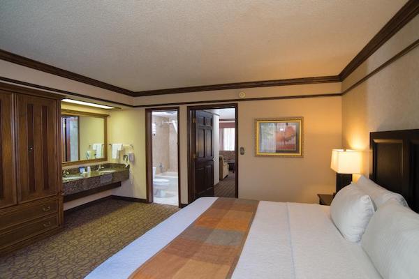 A hotel room looking over a king-size bed. A vanity with dual sinks is seen opposite the bed along with a door that is open to reveal a toilet and bathtub. A second door leads to a living area where seating can be seen. 