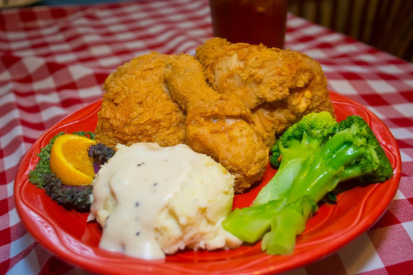 A red plate is seen on the red-and-white-checkered tablecloth. The plate holds three pieces of fried chicken, two broccoli stalks and a scoop of mashed potatoes topped with gravy. A glass of tea is seen in the background. 