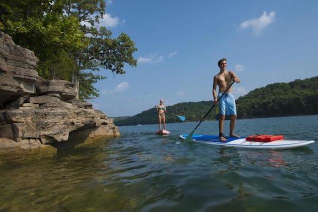 A lake setting. Water fills the frame with a low, rock cliff on one side and wooded tree line on the right. Two paddleboarders are seen on the water. A guy in front and a girl in the back. Both are paddling