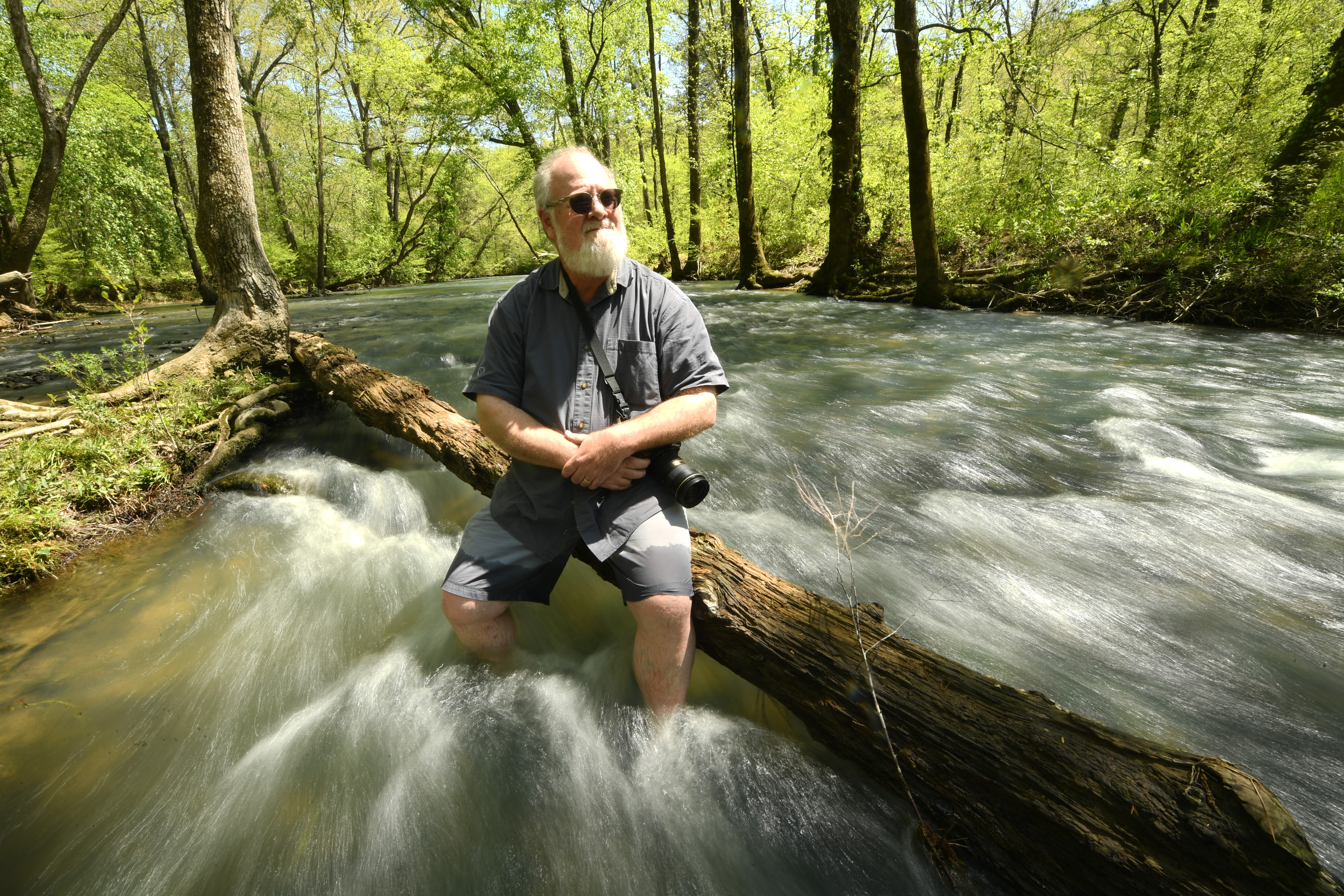 A picture of a man sitting on a fallen log in the middle of rushing water.