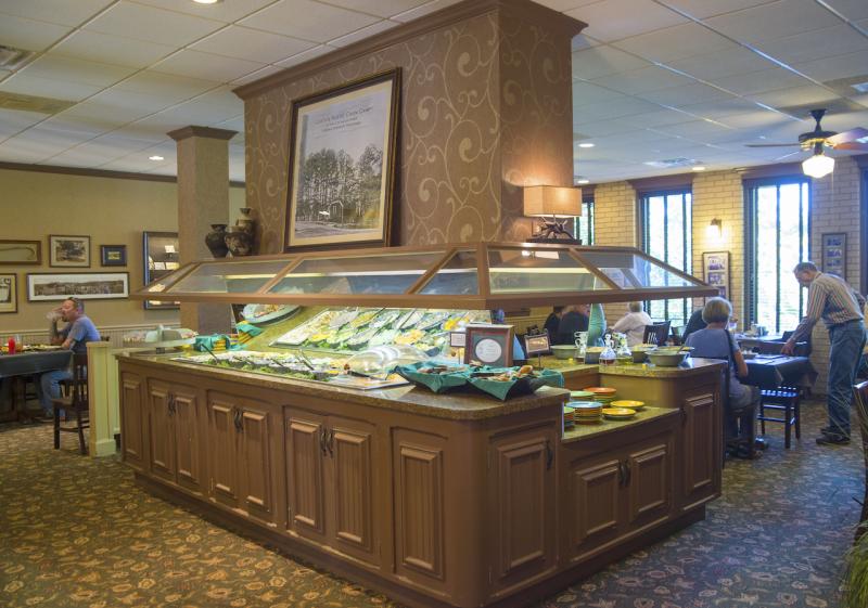 A large restaurant dining space. In the center, cabinetry holds a built-in salad bar with a nose-guard shield over the offerings. Dining tables with people can be seen around the perimeter. 