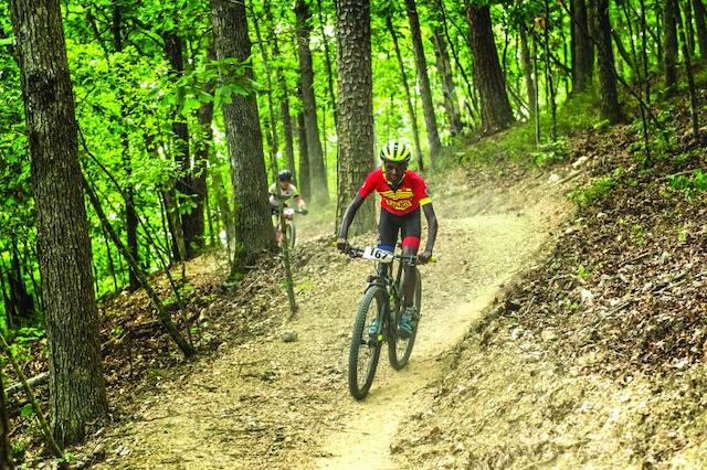 Wooded, hillside setting with a singletrack descending. A mountain biker wearing gear and a helmet is seen on his bike in the front. A second biker can be seen in the background. 
