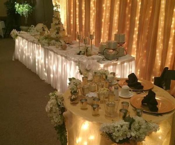A darkened ballroom setting. Two tables are seen close up. One long table is covered with a white tablecloth. Underneath the cloth, strands of lights are illuminated. A round table with two place settings holds votive candles, along with hydrangea flowers. 