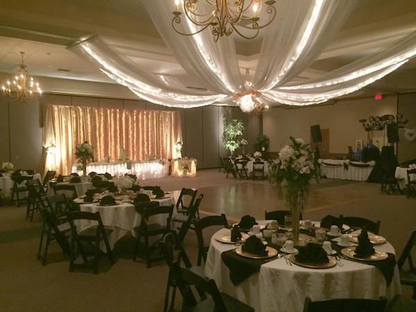 A ballroom. A dance floor is in the center. Round tables covered with tablecloths surround the dancefloor. Each table has eight black chairs. At one end, a long table is illuminated with a strand of lights under a tablecloth. On the ceiling, a piece of draped fabric extends across the room. 