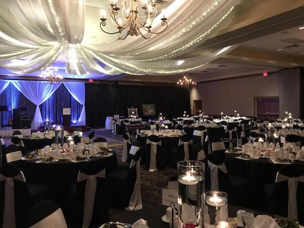 A ballroom. At the front, there is a podium. Black curtain stands line the front of the room to create a softer environment. Round tables are covered with black tablecloths and set for a dinner meal. Each has eight chairs covered with black slipcovers and a cream sash bow. The ceiling is draped with lights and light-colored fabric. 