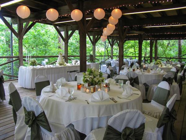 A very large, covered wooden deck area is set up with round tables set for a dinner. The tables are covered with white tablecloths. Each has eight chairs; these chairs are covered with white slipcovers and a green sash. A long table with a cake is seen at one side. Paper lanterns hang from the ceiling. 