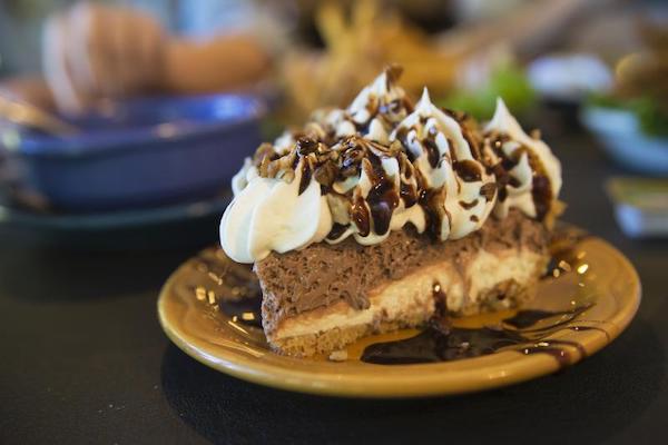 A yellow dessert plate is filled with a slice of pie. The pie layers can be seen from crust to a pudding to a light chocolate filling that is topped with piped whipped cream, chocolate sauce and nuts. 