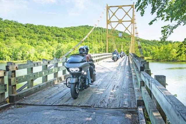 A lake setting. A wooden bridge fills the frame with a motorcycle in front, just crossing over the bridge. 
