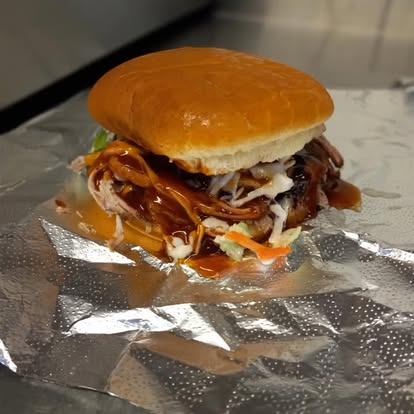 A sandwich topped by a bun has shredded chicken, barbecue sauce and coleslaw sits on a piece of aluminum foil waiting to be wrapped.