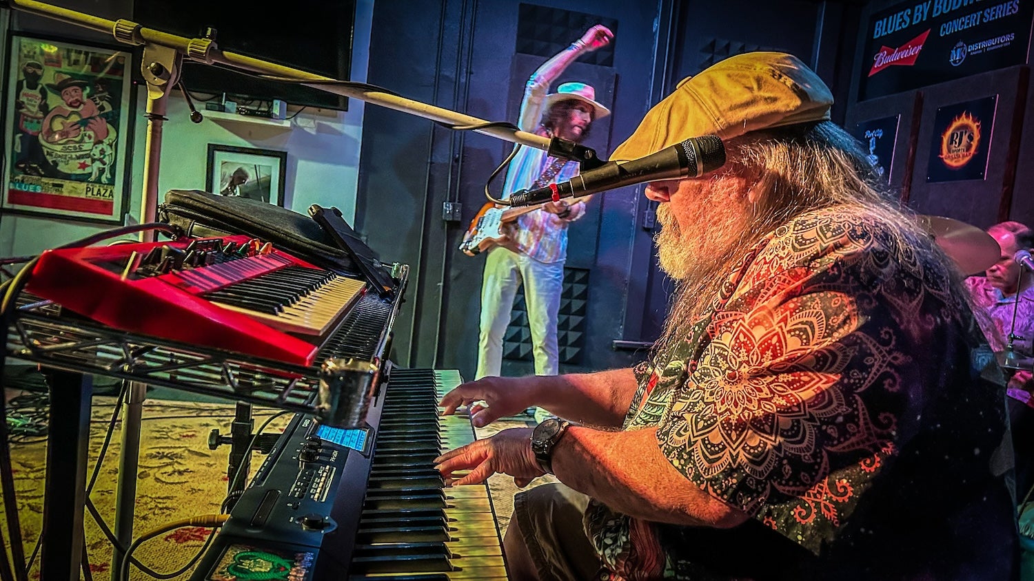 A bearded man plays a keyboard during a live blues performance, with a bandmate playing guitar in the background and vibrant stage lighting illuminating the scene.