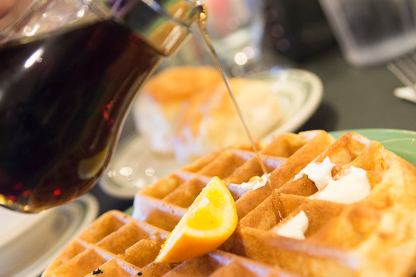 A waffle, topped with butter and an orange slice sits on a plate. A pitcher of syrup is shown being poured over the top. 