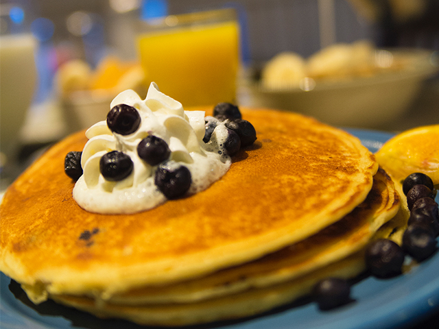 A stack of three golden pancakes sits on a plate. Topped with butter, whipped cream and blueberries. A glass of orange juice is seen in the background. 
