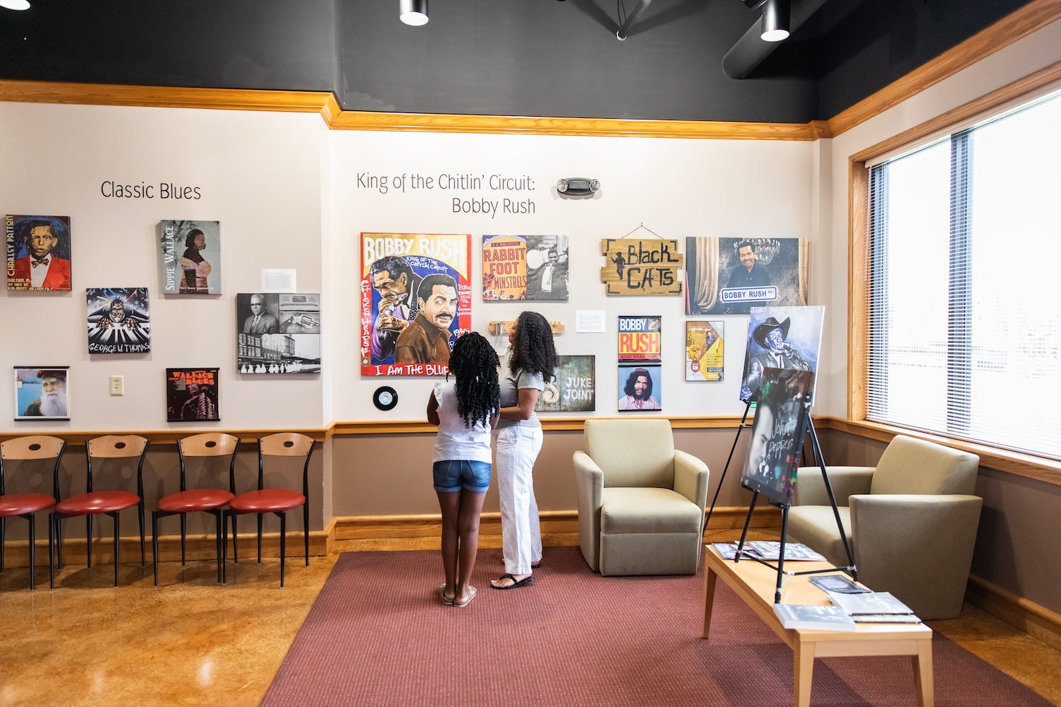 Two women stand in front of a wall exhibit honoring Bobby Rush, with various posters, photos, and signs related to blues music and the Chitlin' Circuit. The display is inside a brightly lit room with chairs, armchairs, and large windows. 