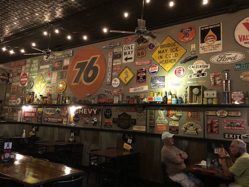 Interior of a retro-style diner with vintage gas station signs and memorabilia covering the walls. Two older men sit and chat at a table beneath string lights and ceiling fans, creating a nostalgic, relaxed setting.