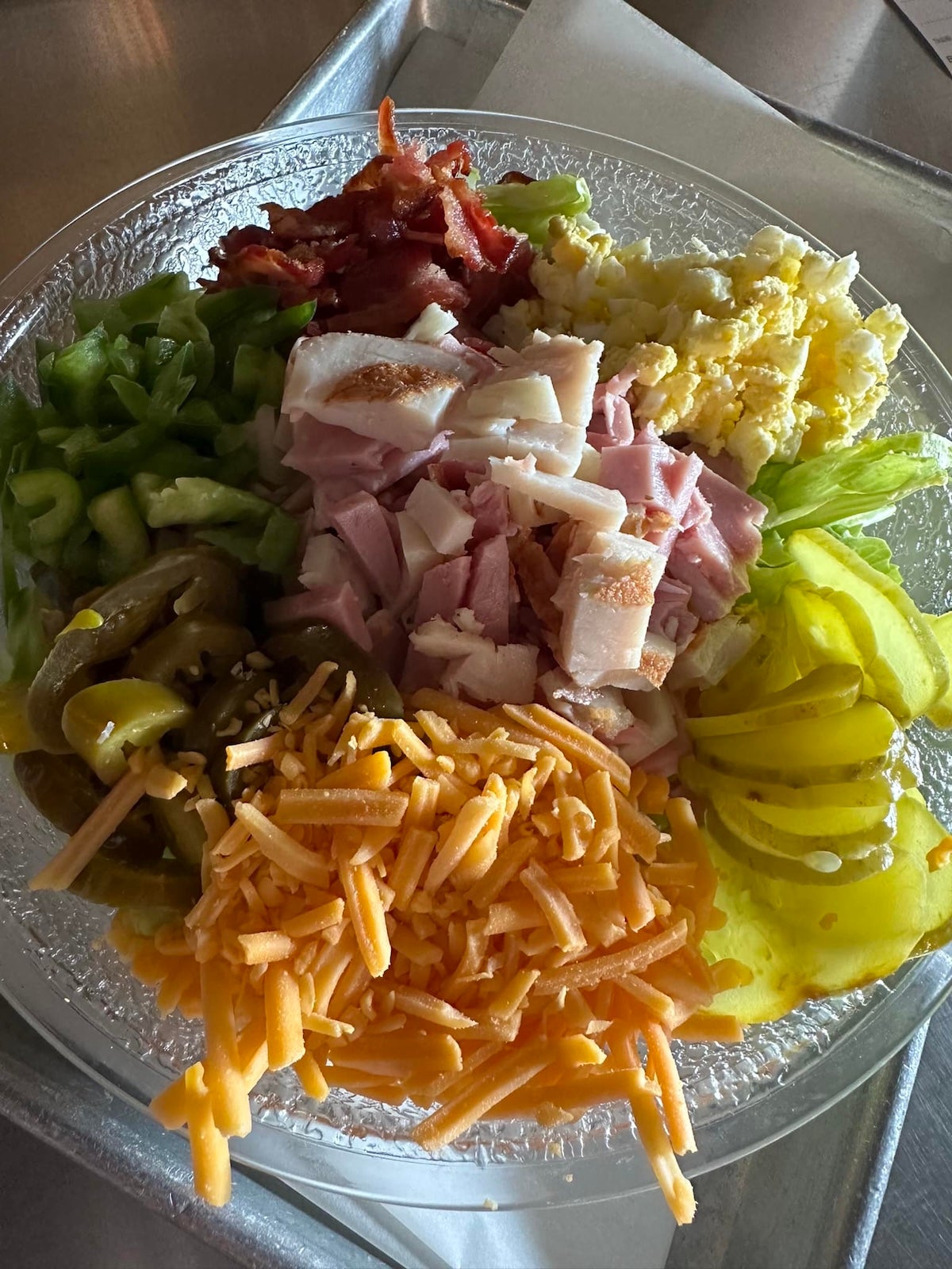 Colorful salad ingredients – chopped green bell peppers, crispy bacon, boiled egg, pickle slices, shredded cheddar cheese and pickled jalapeno peppers – topped with ham and turkey in a bowl on a silver tray.
