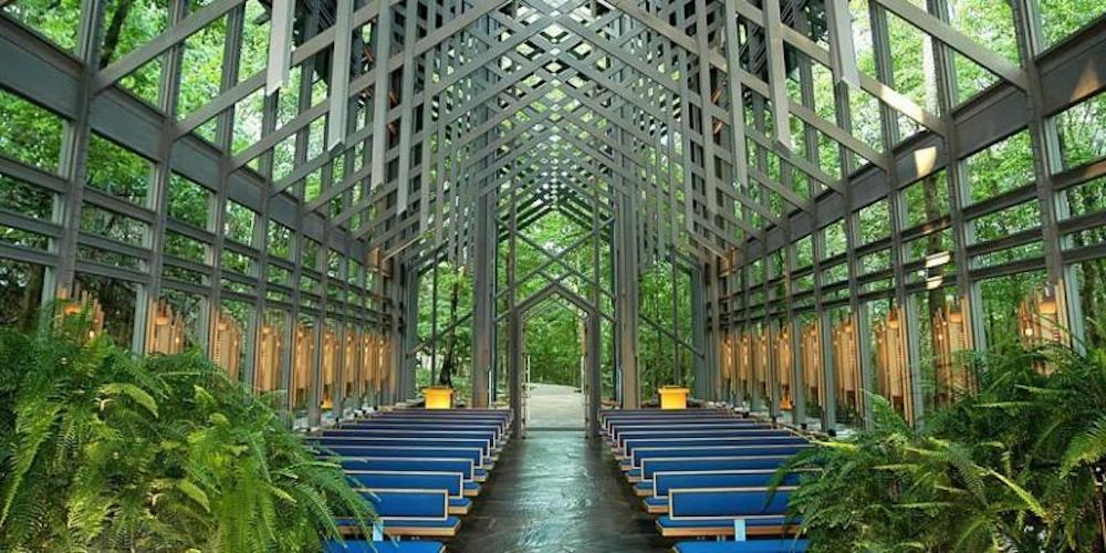 A modern architecture chapel. The sides and ceiling feature a series of crossing beams. Glass sides show a wooded outdoor setting. Sleek, blue pews sit on either side of an aisle. No other accents appear to add to the contemporary, minimalist feel. 