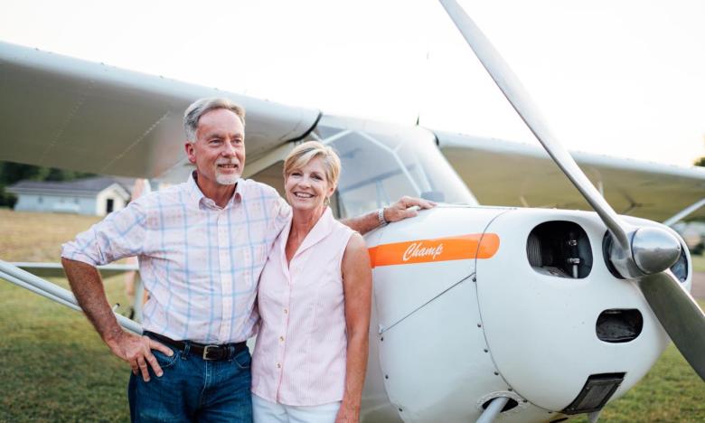 An outdoor setting. A white single-engine plane with an orange stripe takes up the majority of the photography. A middle-aged couple stands near the plane, smiling. Photo source: James Moore.