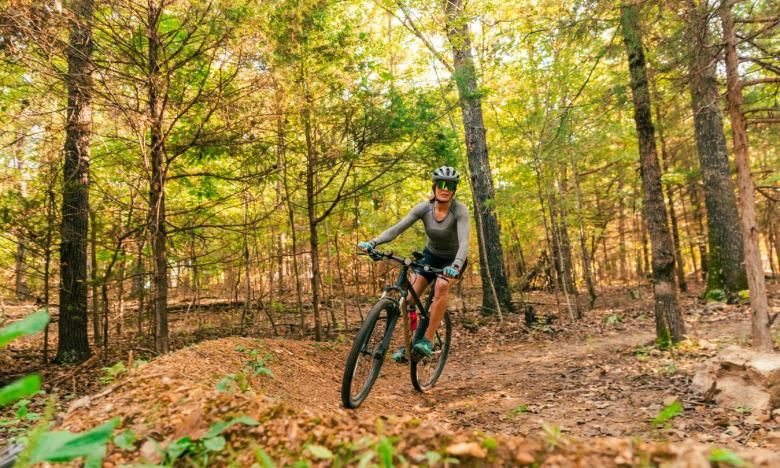 A wooded setting with a mountain bike trail. A woman in workout clothes, a bike helmet and sunglasses rides her bike along the trail. Photo source: Magnetic Creative.