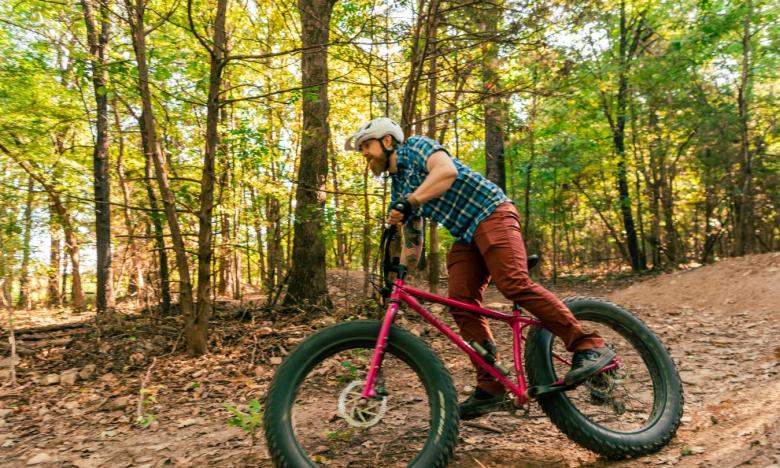 A wooded setting with brown leaves on ground and green leaves on trees. A man is riding a mountain bike. He is wearing a helmet and looking ahead to his path. Photo source: Magnetic Creative.