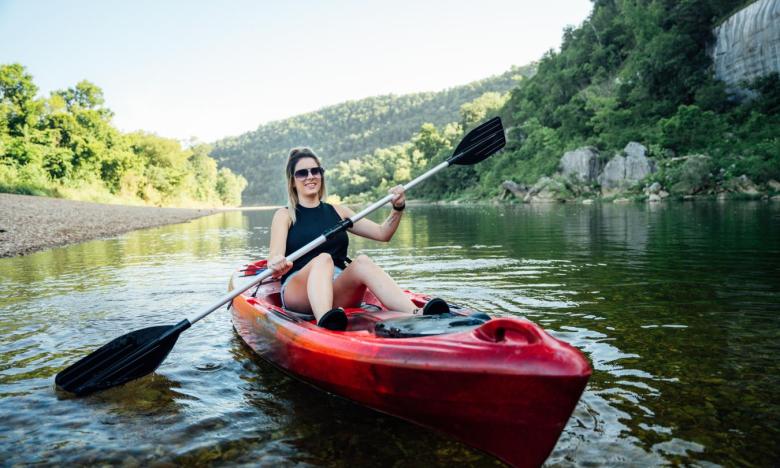The shoreline along the Buffalo National River in Arkansas with green-tree mountains and limestone bluffs in the background. A girl is sitting in a red kayak on the water, holding a paddle dipped into the water on one side. Photo source: James Moore.