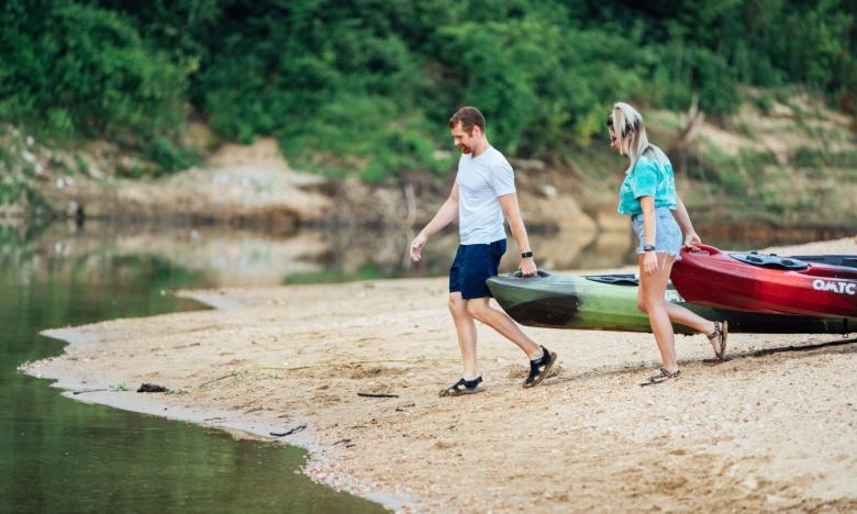 A waterside setting with a shoreline along the Buffalo National River. A man and a woman are each dragging their own kayaks from the shore into the water. They are dressed in shorts and T-shirts. Photo source: James Moore.