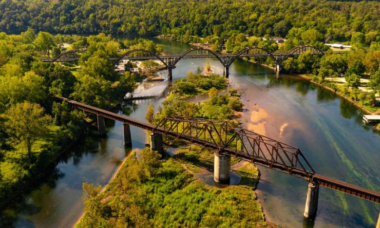 A photograph of an outdoor setting with a body of water near Mountain Home, AR. Shorelines with trees are on either side of the water and an island with trees can be seen in the center. Two bridges traverse the water. A brown, rusted train bridge on concrete pillars is in front. A bridge for vehicle traffic is in the back. Photo source: James Moore.