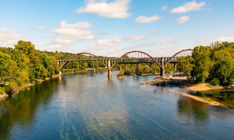 A photograph of an outdoor setting with a body of water near Mountain Home, AR. Shorelines with trees are on either side of the water. A blue sky overhead. An historic-looking bridge with a series of arches leads from one side to the other. Photo source: James Moore.