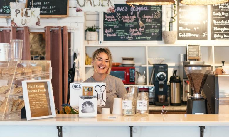 The order bar at Cove Coffee in Mountain Home, Arkansas. Coffee making machines are seen on front and back counters. Boards with handwritten menus line the walls. A case of muffins and pastries sits on the counter. A girl smiles behind the order screen. Photo source: Magnetic Creative.
