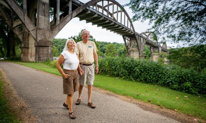 An outdoor setting along a waterway with Cotter Bridge crossing the White River in the background. A paved trail, known as the Big Spring Nature Trail, is seen in the foreground. Green trees, grass and bushes line the shore. An arched bridge is seen in the background. An older couple walks hand-in-hand on the paved portion of the trail. Photo source: James Moore.