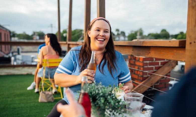 The rooftop of Rio Burrito. Two people are sitting at a small bistro table. You can see the face of only one, a woman in a blue T-shirt. She is smiling and holding a beverage that is in a glass bottle. A second table with a couple is seen in the background. Photo source: James Moore.