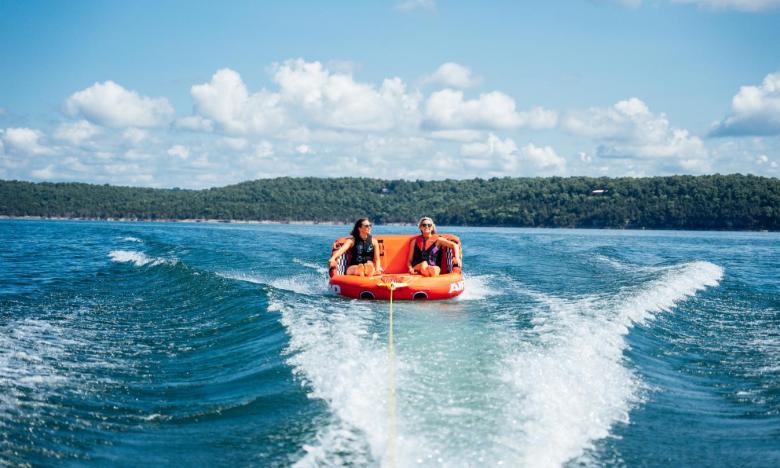 A sunny, outdoor setting on Norfork Lake near Mountain Home, Arkansas. Inside the wake behind a boat, two women are seen on a large inner tube. Trees and blue lake water are seen in the background. Photo source: James Moore.
