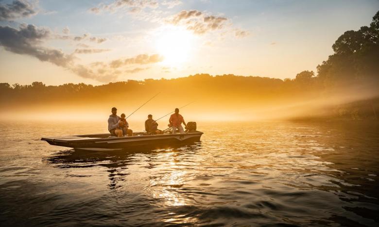 An outdoor setting on the waters of the White River. The sun is rising and the fog is beginning to lift from the water. A fishing boat holds a guide along with a father and two young sons. The father and one of the sons each have a fishing pole in the water. Photo source: James Moore.