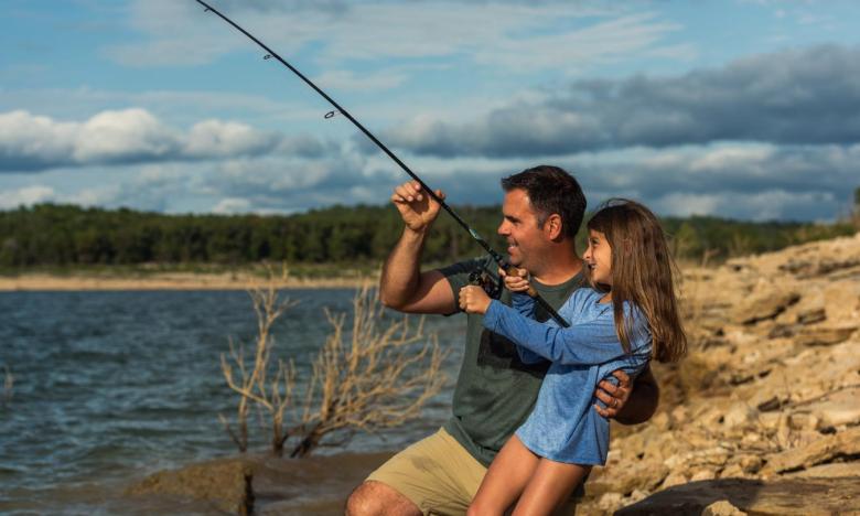 An outdoor setting near the White River in Mountain Home, Arkansas. It is a sunny day with white clouds in a blue sky. A rocky bank is seen next to the water. A young girl holds a fishing pole and appears to be reeling in a catch. Her father kneels beside her, steadying her with one hand and the pole with his other. Photo source: Magnetic Creative.