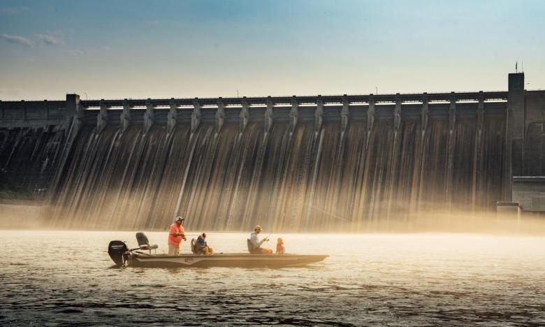On the water of the White River near the Bull Shoals dam at Mountain Home. The dam is seen in the background and a blue sky is overhead. A fishing boat is in the foreground. A guide from Gaston’s White River Resort and a father each hold fishing poles that have been cast into the water. Two young boys wearing life jackets sit inside the boat. Photo source: James Moore.