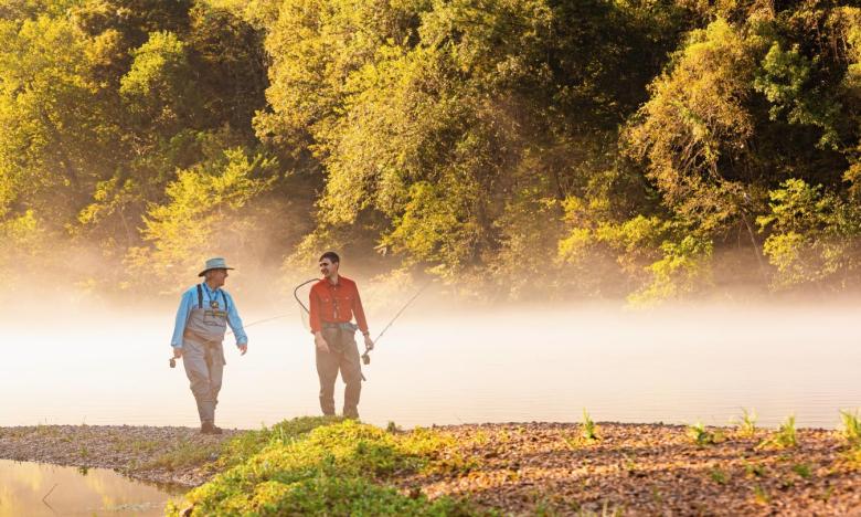 An outdoor setting along a waterway, possibly the White River near Mountain Home, Arkansas. The time of day is likely early morning as a fog is rising off the water. Two men, one older and one younger, are seen walking along the bank. Both wear waders and hold a rod and reel. The younger man also has a fishing net. Photo source: Magnetic Creative.