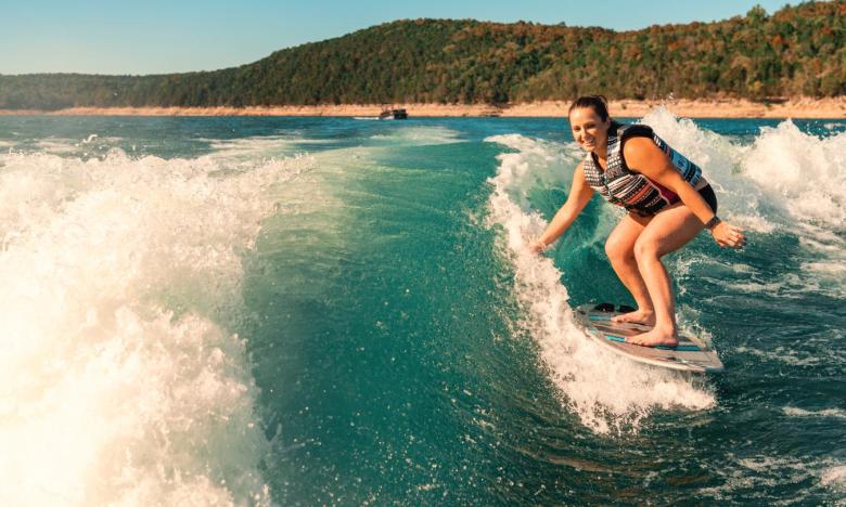 A lake setting near Mountain Home, Arkansas. Blue sky and green-tree hills are seen in the background. Blue water and white wake fill the rest of the photograph. A girl is seen on a board surfing in the wake. Photo source: Magnetic Creative.