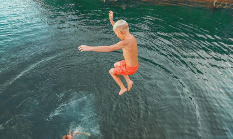 An outdoor setting on a lake near Mountain Home, Arkansas. The photograph is taken from overhead looking into the lake water. A young boy with blonde hair wearing orange swim trunks is seen jumping into the water. His arms are in the air and his legs are tucked in a chair position. Photo source: Magnetic Creative.