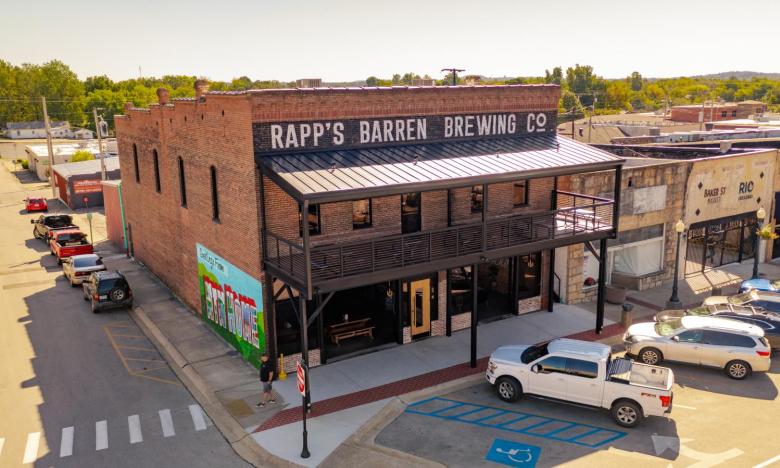 A downtown setting in Mountain Home, Arkansas. A restored building houses a brewery and restaurant. The sign painted across the top of the building reads Rapp’s Barren Brewing Co. An outdoor, covered patio is seen on the second floor. Cars and trucks are parked along the front of the building’s downtown Main Street-style setting. A colorful mural that reads Greetings from Mtn Home is seen on the side of the building that is not attached to the rest of the downtown district buildings. Photo source: James Moo
