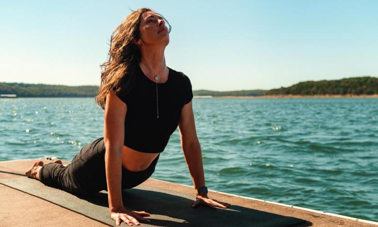 A lake setting near Mountain Home, Arkansas. Water, shoreline and green hills are seen in the background. In the foreground a woman is on a yoga mat on a dock. She is dressed in black yoga pants and a black cropped top with a Y necklace. She is in Cobra pose, posturing toward the sun. Photo source: Magnetic Creative.
