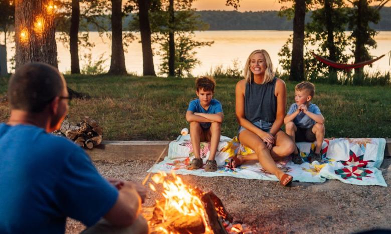 An outdoor setting along the banks of a waterway. The area nearest the water is grassy. In the foreground a family is on a camping pad. A mom and two boys sit on a colorful quilt near a campfire. A dad sits on the opposite side of the fire with his back to the camera. He holds a skewer with a marshmallow on one end. A box of graham crackers and a bag of marshmallows sit on the ground next to him. The time is near sunset. A hammock hangs in one tree seen by the water. Photo source: James Moore.