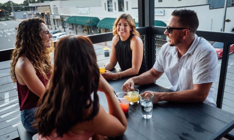 An outdoor rooftop setting at Rapp’s Brewing in Mountain Home, Arkansas. There is a blue sky in the background and the space’s railing can be seen around the edge. Four people are seated at a picnic table; three women and a man. They are talking and enjoying drinks. Photo source: James Moore.