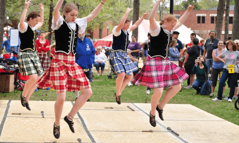 Four young women in traditional Scottish attire are doing a Highland dance demonstration for a crowd of onlookers. All four dancers have both feet off the ground and hands in the air.