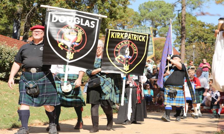 A parade of people are dressed in traditional Scottish Highland dress—tartan kilts, black brogues and long socks, red bonnets with crest badges, sporrans with chains. Some are holding banners displaying Scottish Clans, featuring names and crests. The “Douglas” and “Kirkpatrick” Clan banners are visible. Another man is carrying a flag.